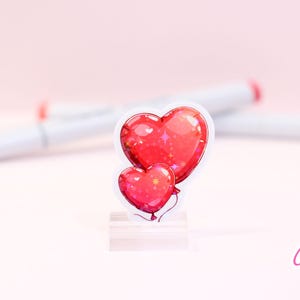 Peut inclure: Un autocollant repr&eacute;sentant deux ballons en forme de c&oelig;ur rouges avec un effet scintillant. L'autocollant a une bordure blanche et est pr&eacute;sent&eacute; sur un support transparent. L'arri&egrave;re-plan comprend des marqueurs blancs avec des pointes rouges et le texte "Cypriana Stickers".