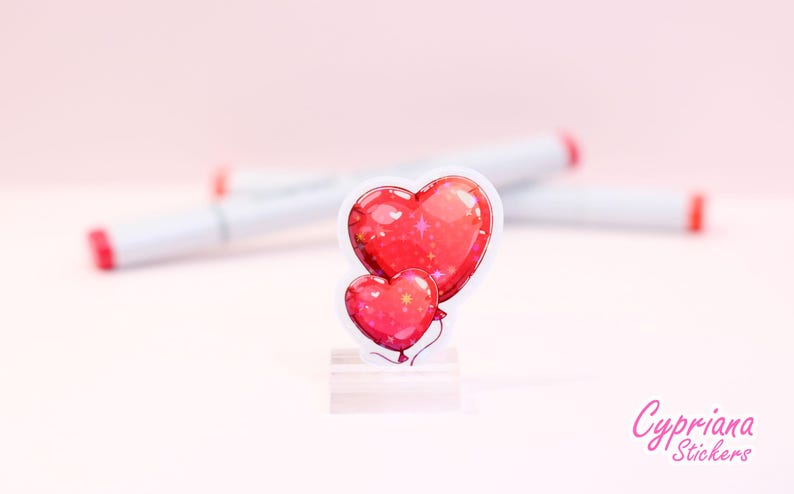 Peut inclure: Un autocollant repr&eacute;sentant deux ballons en forme de c&oelig;ur rouges avec un effet scintillant. L'autocollant a une bordure blanche et est pr&eacute;sent&eacute; sur un support transparent. L'arri&egrave;re-plan comprend des marqueurs blancs avec des pointes rouges et le texte "Cypriana Stickers".