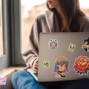 Pode incluir: Um computador port&aacute;til com v&aacute;rios adesivos coloridos na tampa. Os adesivos apresentam um sapo de desenho animado, um gato comendo pizza, uma flor e uma menina com cabelo azul e rosa. O laptop est&aacute; aberto e a tela &eacute; vis&iacute;vel.