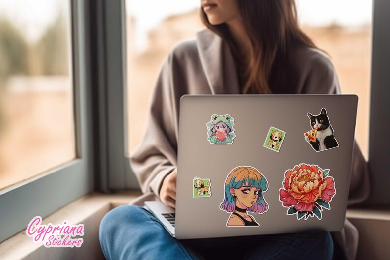 Pode incluir: Um computador port&aacute;til com v&aacute;rios adesivos coloridos na tampa. Os adesivos apresentam um sapo de desenho animado, um gato comendo pizza, uma flor e uma menina com cabelo azul e rosa. O laptop est&aacute; aberto e a tela &eacute; vis&iacute;vel.