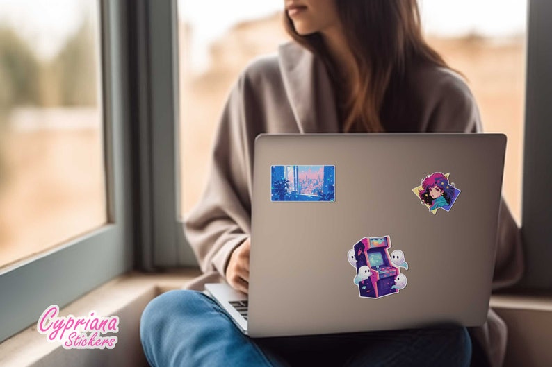 Puede incluir: Un port&aacute;til plateado con tres pegatinas en la tapa. Las pegatinas son de un juego de arcade retro, una ciudad y una chica de dibujos animados. El port&aacute;til est&aacute; sentado en el regazo de una persona que lleva pantalones vaqueros azules y un su&eacute;ter beige.
