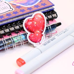 Peut inclure: Un autocollant repr&eacute;sentant deux ballons rouges en forme de c&oelig;ur avec des d&eacute;tails blancs, plac&eacute; pr&egrave;s d'une pile de carnets et de deux marqueurs blancs avec des pointes rouges et roses. Les carnets ont des reliures &agrave; spirale et le premier porte l'inscription "I READ PAST MY BEDTIME".