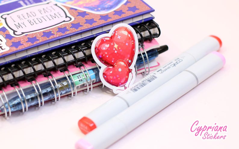Peut inclure: Un autocollant repr&eacute;sentant deux ballons rouges en forme de c&oelig;ur avec des d&eacute;tails blancs, plac&eacute; pr&egrave;s d'une pile de carnets et de deux marqueurs blancs avec des pointes rouges et roses. Les carnets ont des reliures &agrave; spirale et le premier porte l'inscription "I READ PAST MY BEDTIME".