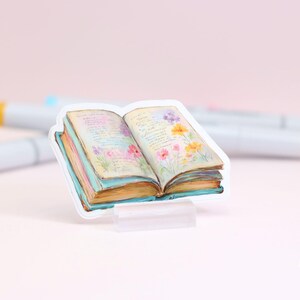 May include: A sticker of an open book with watercolor illustrations of flowers and text. The book has a light blue cover and pages with a vintage look. The sticker is on a clear stand, with markers in the background.