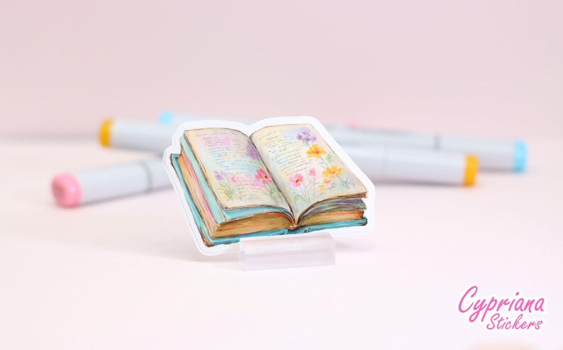 May include: A sticker of an open book with watercolor illustrations of flowers and text. The book has a light blue cover and pages with a vintage look. The sticker is on a clear stand, with markers in the background.