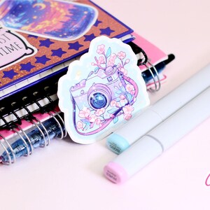 May include: A sticker featuring a camera design with floral accents, resting on a stack of notebooks and two light gray markers. The sticker has a light blue border and the camera is decorated with pink flowers. The notebooks have spiral bindings.