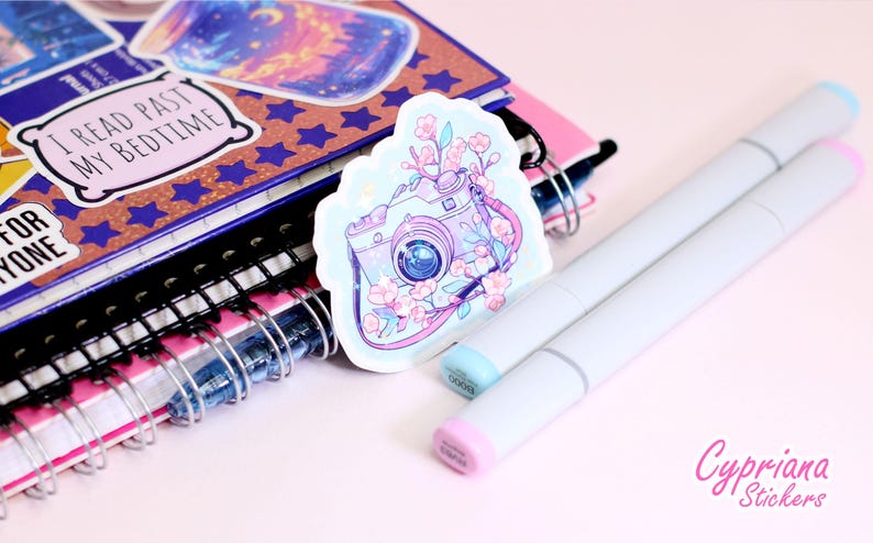 May include: A sticker featuring a camera design with floral accents, resting on a stack of notebooks and two light gray markers. The sticker has a light blue border and the camera is decorated with pink flowers. The notebooks have spiral bindings.