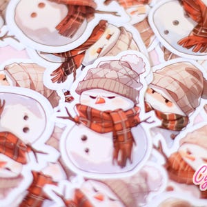 May include: A collection of snowman stickers with various poses and expressions. The snowmen are wearing brown and red plaid scarves and hats. Some snowmen have a stick for arms and a carrot nose.