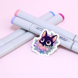 May include: A black cat sticker with large eyes surrounded by purple, pink and blue flowers. The sticker is on a pink surface with four Copic markers in the background.