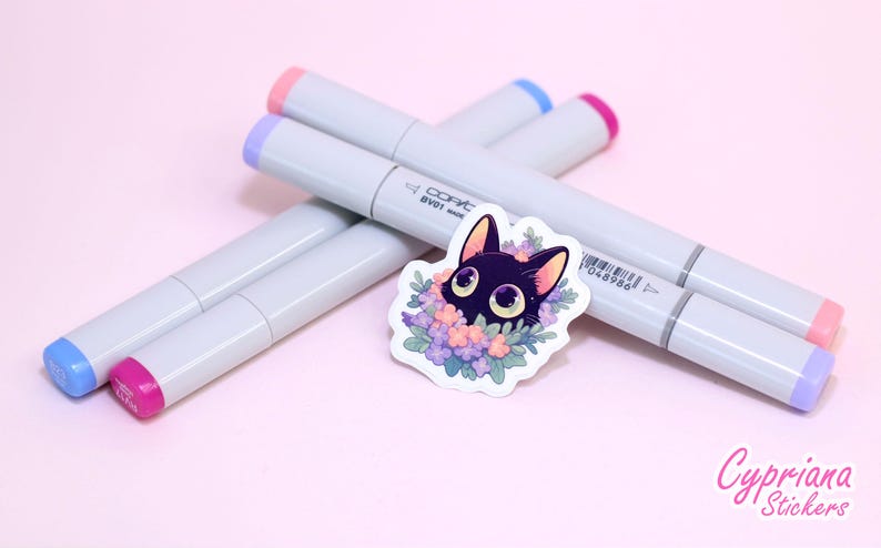 May include: A black cat sticker with large eyes surrounded by purple, pink and blue flowers. The sticker is on a pink surface with four Copic markers in the background.