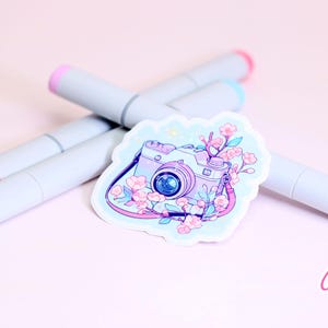 May include: A sticker featuring a camera design with floral accents in shades of pink and purple. The sticker is surrounded by four gray markers with pink and blue tips, set against a soft pink background. The sticker has the text "Cypriana Stickers".