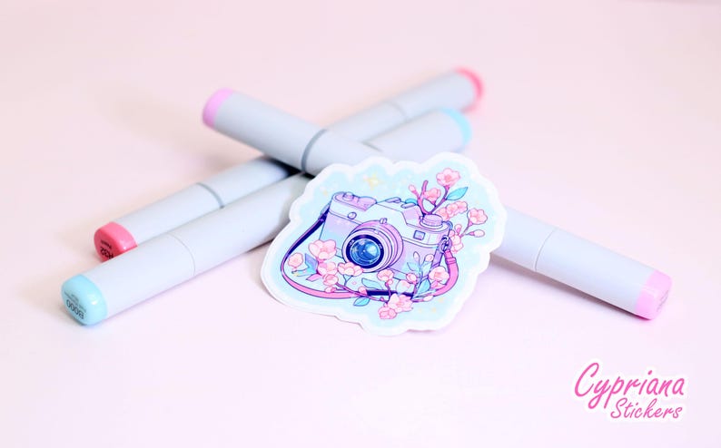 May include: A sticker featuring a camera design with floral accents in shades of pink and purple. The sticker is surrounded by four gray markers with pink and blue tips, set against a soft pink background. The sticker has the text "Cypriana Stickers".