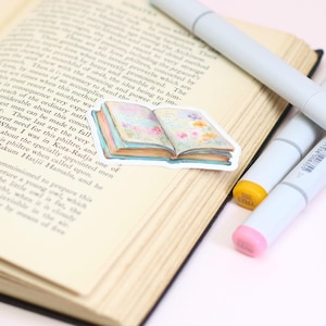 May include: An open book with text, featuring a sticker of a watercolor-style book with floral accents. Four gray markers with colorful tips are arranged nearby. The sticker and markers are on a light-colored surface.