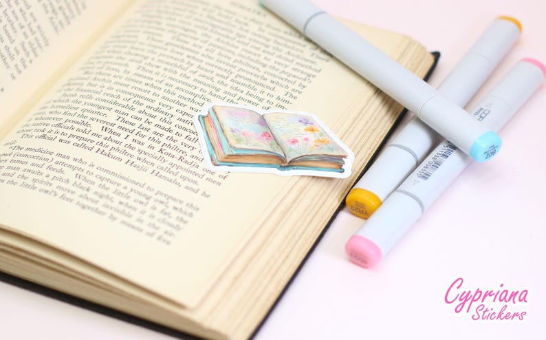 May include: An open book with text, featuring a sticker of a watercolor-style book with floral accents. Four gray markers with colorful tips are arranged nearby. The sticker and markers are on a light-colored surface.