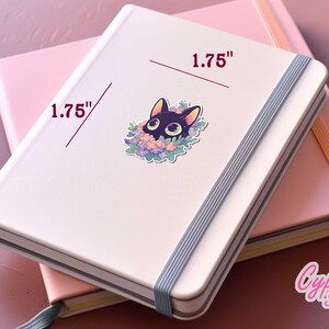 May include: A white notebook with a black cat sticker on the front. The sticker is 1.75 inches wide and features a black cat with green eyes peeking out from a wreath of purple and pink flowers. The notebook is closed and has a grey elastic band.
