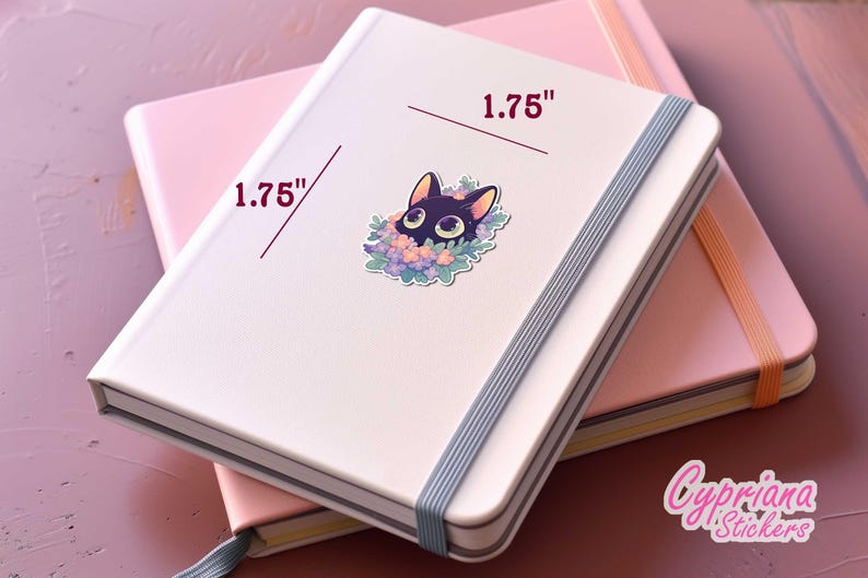 May include: A white notebook with a black cat sticker on the front. The sticker is 1.75 inches wide and features a black cat with green eyes peeking out from a wreath of purple and pink flowers. The notebook is closed and has a grey elastic band.