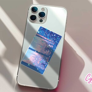 May include: A white iPhone with a blue and pink sticker on the back. The sticker features a cityscape with stars and a pink sky. The text "Cypriana Stickers" is printed in pink on a white background.