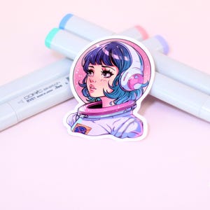 May include: A sticker featuring a colorful illustration of a girl with blue hair wearing a space suit and headphones. The background is pink with stars. The sticker is on a white surface with markers.