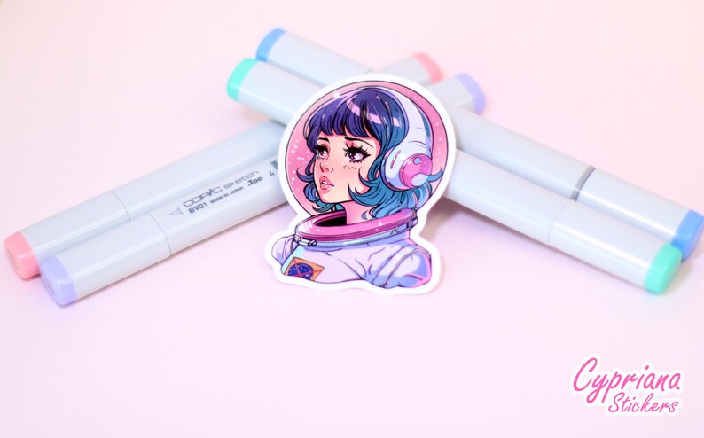 May include: A sticker featuring a colorful illustration of a girl with blue hair wearing a space suit and headphones. The background is pink with stars. The sticker is on a white surface with markers.