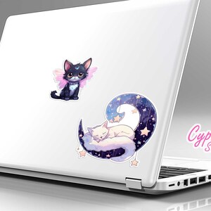 May include: Two cute cat stickers on a white laptop. One sticker features a black cat with pink wings and the other sticker features a white cat sleeping on a crescent moon with stars.