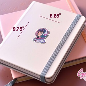 May include: A white notebook with a blue ribbon closure and a sticker of a girl in a space suit. The notebook is 2.75 inches wide and 2.25 inches tall. The sticker is from Cypriana Stickers.