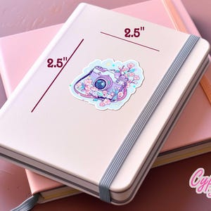 May include: A light pink notebook with a 2.5-inch sticker of a purple camera surrounded by flowers. The sticker is on top of another pink notebook. The notebooks are on a pink surface. The sticker is from Cypriana Stickers.