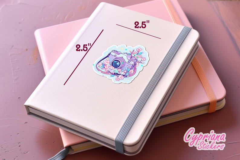 May include: A light pink notebook with a 2.5-inch sticker of a purple camera surrounded by flowers. The sticker is on top of another pink notebook. The notebooks are on a pink surface. The sticker is from Cypriana Stickers.