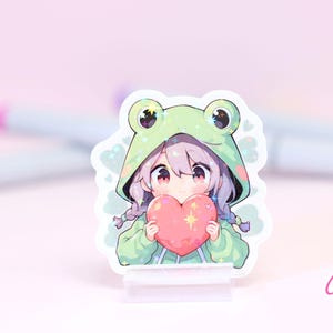Pode incluir: Um adesivo com um personagem de estilo anime com cabelo roxo usando um moletom verde de sapo e segurando um cora&ccedil;&atilde;o rosa. O personagem tem uma express&atilde;o fofa e est&aacute; cercado por brilho. O adesivo est&aacute; em um suporte de acr&iacute;lico transparente.