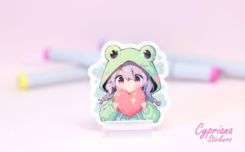 Pode incluir: Um adesivo com um personagem de estilo anime com cabelo roxo usando um moletom verde de sapo e segurando um cora&ccedil;&atilde;o rosa. O personagem tem uma express&atilde;o fofa e est&aacute; cercado por brilho. O adesivo est&aacute; em um suporte de acr&iacute;lico transparente.