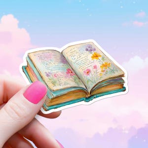 May include: A sticker of an open book with watercolor illustrations of flowers and handwritten text. The book has a teal cover and gold-colored pages. Pink, yellow, and purple flowers are depicted on the pages. The sticker is held by a hand with pink nail polish.