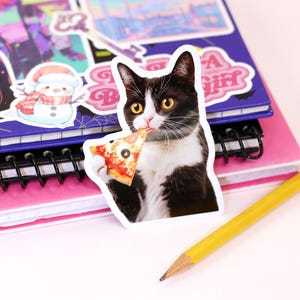 以下が含まれることがあります： ピザを食べている白黒の猫のステッカーが、他のステッカーが付いたピンクと紫のノートに貼られています。黄色の鉛筆がノートの横にあります。ステッカーには白い縁があります。「Cypriana Stickers」の文字が右下にあります。