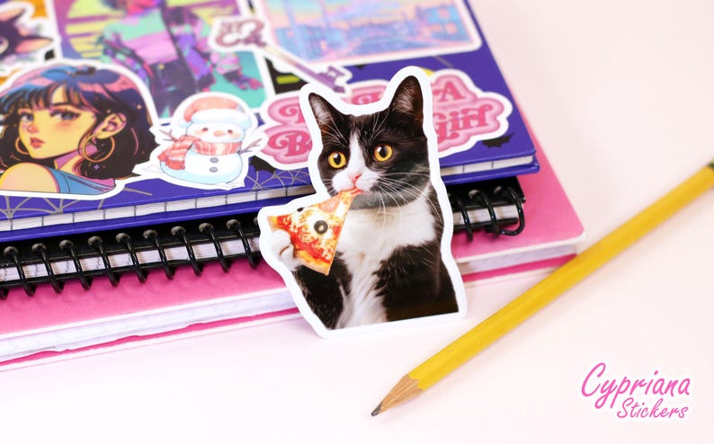 以下が含まれることがあります： ピザを食べている白黒の猫のステッカーが、他のステッカーが付いたピンクと紫のノートに貼られています。黄色の鉛筆がノートの横にあります。ステッカーには白い縁があります。「Cypriana Stickers」の文字が右下にあります。