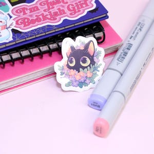 May include: A black cat sticker with large green eyes surrounded by purple and pink flowers. The sticker is on a pink and white notebook with a black spiral binding. Two grey markers are also in the image.