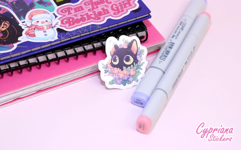 May include: A black cat sticker with large green eyes surrounded by purple and pink flowers. The sticker is on a pink and white notebook with a black spiral binding. Two grey markers are also in the image.