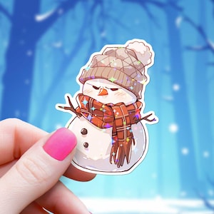 May include: A sticker featuring a cartoon snowman with an angry expression. The snowman wears a brown knit hat with a white pom-pom, a plaid scarf, and twig arms. The sticker has a holographic, glittery effect.