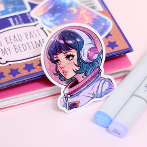 May include: A sticker featuring a colorful illustration of a girl wearing an astronaut suit and a headset. The girl has blue hair and is looking to the side. The sticker is on a pink surface with other stickers and markers.