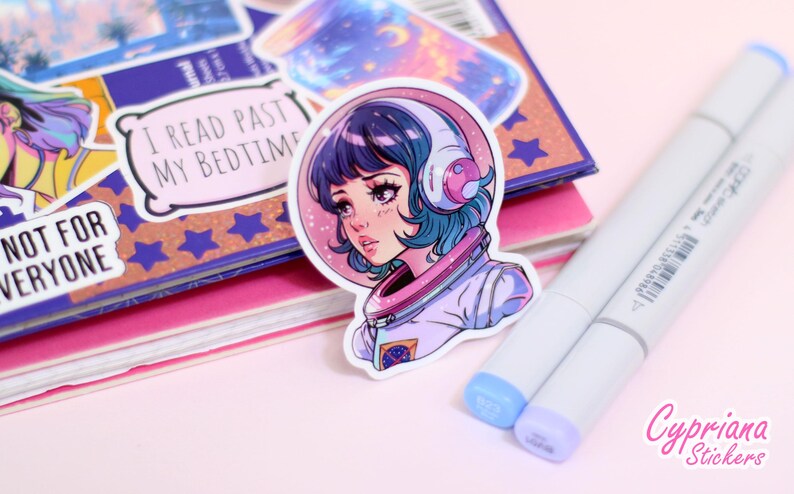 May include: A sticker featuring a colorful illustration of a girl wearing an astronaut suit and a headset. The girl has blue hair and is looking to the side. The sticker is on a pink surface with other stickers and markers.