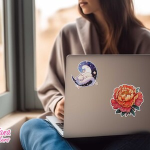 May include: A silver laptop with two stickers. One sticker features a white cat sleeping on a crescent moon with stars. The other sticker is a red and yellow peony flower.