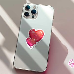 Peut inclure: Un autocollant repr&eacute;sentant deux ballons en forme de c&oelig;ur rouges avec une finition brillante et des contours blancs, coll&eacute; au dos d'un smartphone argent&eacute;. L'autocollant a un effet scintillant. La marque "Cypriana Stickers" est visible.