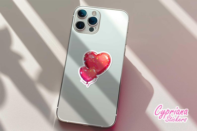 Peut inclure: Un autocollant repr&eacute;sentant deux ballons en forme de c&oelig;ur rouges avec une finition brillante et des contours blancs, coll&eacute; au dos d'un smartphone argent&eacute;. L'autocollant a un effet scintillant. La marque "Cypriana Stickers" est visible.
