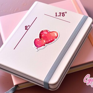 Peut inclure: Un carnet blanc avec un autocollant de deux coeurs rouges. L'autocollant est sur un carnet rose. L'autocollant mesure 4,4 cm de large. Le carnet a une bande &eacute;lastique grise.
