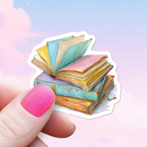 Puede incluir: Una pegatina que representa una pila de libros coloridos. Los libros tienen páginas abiertas con texto visible, y las cubiertas son en tonos de azul, naranja y morado. La pegatina la sostiene una persona con esmalte de uñas rosa.