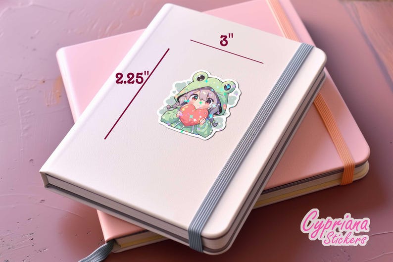 Pode incluir: Um adesivo com uma menina de desenho animado vestindo um moletom verde de sapo e segurando um cora&ccedil;&atilde;o rosa. O adesivo tem 7,6 cm de altura e 5,7 cm de largura. O adesivo est&aacute; em um caderno rosa e branco.