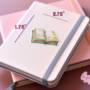 May include: A sticker of an open book with floral illustrations, measuring 2.75 inches wide and 1.75 inches tall, is placed on a white notebook. The notebook is on top of a pink notebook. The sticker has a watercolor style.