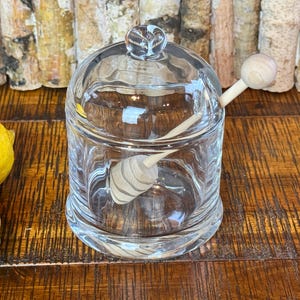 May include: A clear glass jar with a lid and two wooden honey dippers. The jar sits on a wooden surface with two yellow lemons in the background. The jar is designed to hold honey or other condiments.