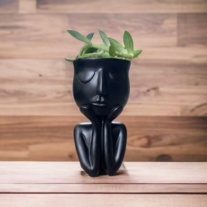 May include: A black ceramic planter shaped like a face, with closed eyes and hands supporting the chin. The planter holds a small green succulent. The background is a wooden surface.