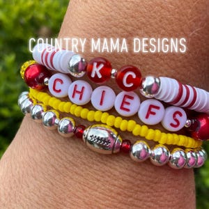Könnte beinhalten: Ein Stapel Armbänder mit roten, weißen und gelben Perlen. Die Armbänder buchstabieren "KC Chiefs" mit einem Fußball-Anhänger.