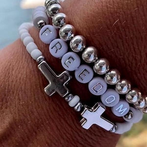 May include: Three stacked bracelets with white and silver beads. One bracelet spells out the word "FREEDOM" with letter beads. Two bracelets have silver cross charms. The text "Ozark Mama Designs" is in the upper left corner.