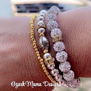 Könnte beinhalten: Eine Auswahl an Armbändern mit Perlen in Gold, Silber und Kristall. Ein Armband hat eine fußballförmige Perle. Der Text "Ozark Mama Designs" ist sichtbar.
