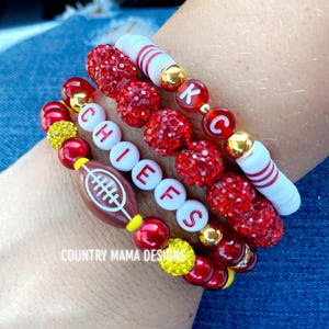 Könnte beinhalten: Ein Set aus sechs roten, weißen und gelben Perlenarmbändern mit dem Wort "CHIEFS" in weißen Perlen und den Buchstaben "KC" in roten Perlen. Die Armbänder haben außerdem einen Fußball-Anhänger.